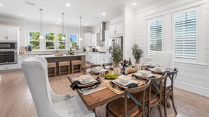 A modern kitchen with white cabinetry, a central island, and a dining area with a wooden table and chairs. - Luxury Prominence Home – Pool, Beach & Golf Cart (Inlet Beach)