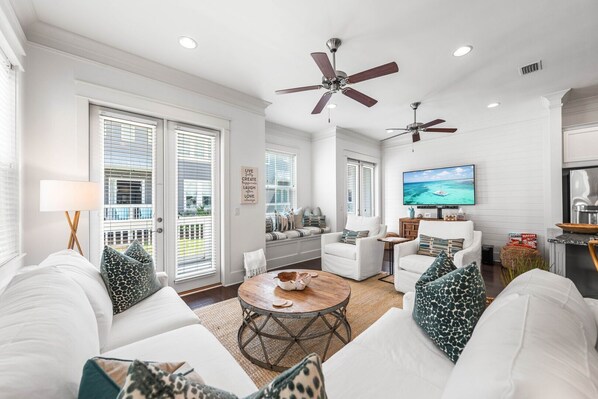 A spacious living room with white furniture, a ceiling fan, a TV, and large windows with blinds. - 30A Coastal Therapy – Pool, Beach & Golf Cart (Inlet Beach)