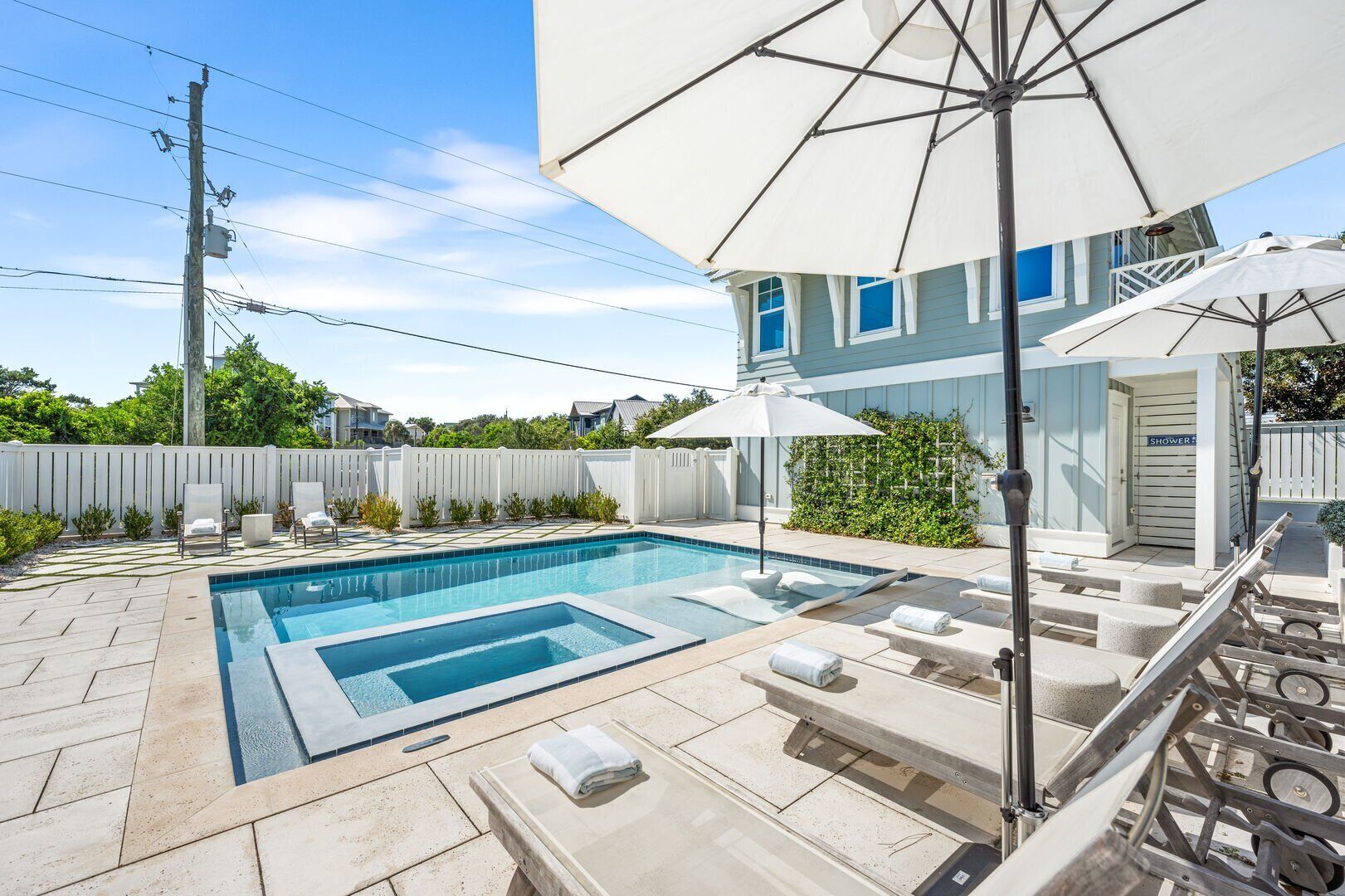 Ein Pool mit Liegestühlen und Sonnenschirmen, ein weißer Zaun und ein Haus mit einer blauen Tür, auf der "SHOWER" steht.
