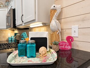A kitchen counter with a tray holding blue glass jars, a white towel, a wooden bird sculpture, a small succulent plant, and a pink sign with text. - Private Heated Pool, Pet Friendly, Golf Cart Included (Port Aransas)