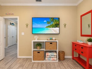 A room with a television displaying a beach scene, a white console table with baskets, a red vanity, and a hallway leading to a door. - Private Heated Pool, Pet Friendly, Golf Cart Included (Port Aransas)