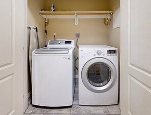 A laundry room with a front-loading washer and dryer, a shelf above the dryer, and a mop on the wall. - Private Heated Pool, Pet Friendly, Golf Cart Included (Port Aransas)