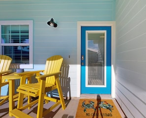 A room with a blue door, two yellow Adirondack chairs, a small table, and a doormat with crab illustrations. - Private Heated Pool, Pet Friendly, Golf Cart Included (Port Aransas)