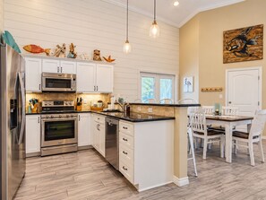 A modern kitchen with white cabinetry, stainless steel appliances, and a dining area with white chairs. - Private Heated Pool, Pet Friendly, Golf Cart Included (Port Aransas)