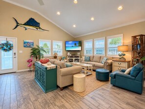 A spacious living room with a large TV, comfortable seating, and a view of the outdoors through large windows. - Private Heated Pool, Pet Friendly, Golf Cart Included (Port Aransas)