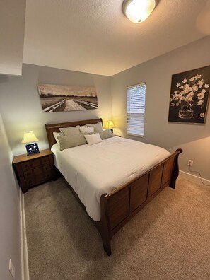 Quarto com cama de madeira, duas mesas de cabeceira com abajures, um quadro na parede e uma janela com persianas.