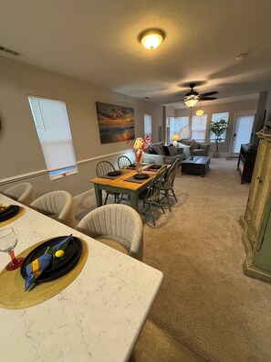Uma sala de jantar com uma mesa de mármore, cadeiras e um vaso com flores vermelhas. Há um quadro na parede e um ventilador de teto.