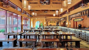 Un restaurante con mesas y bancos de madera, una zona de bar y un techo alto con luces colgantes.