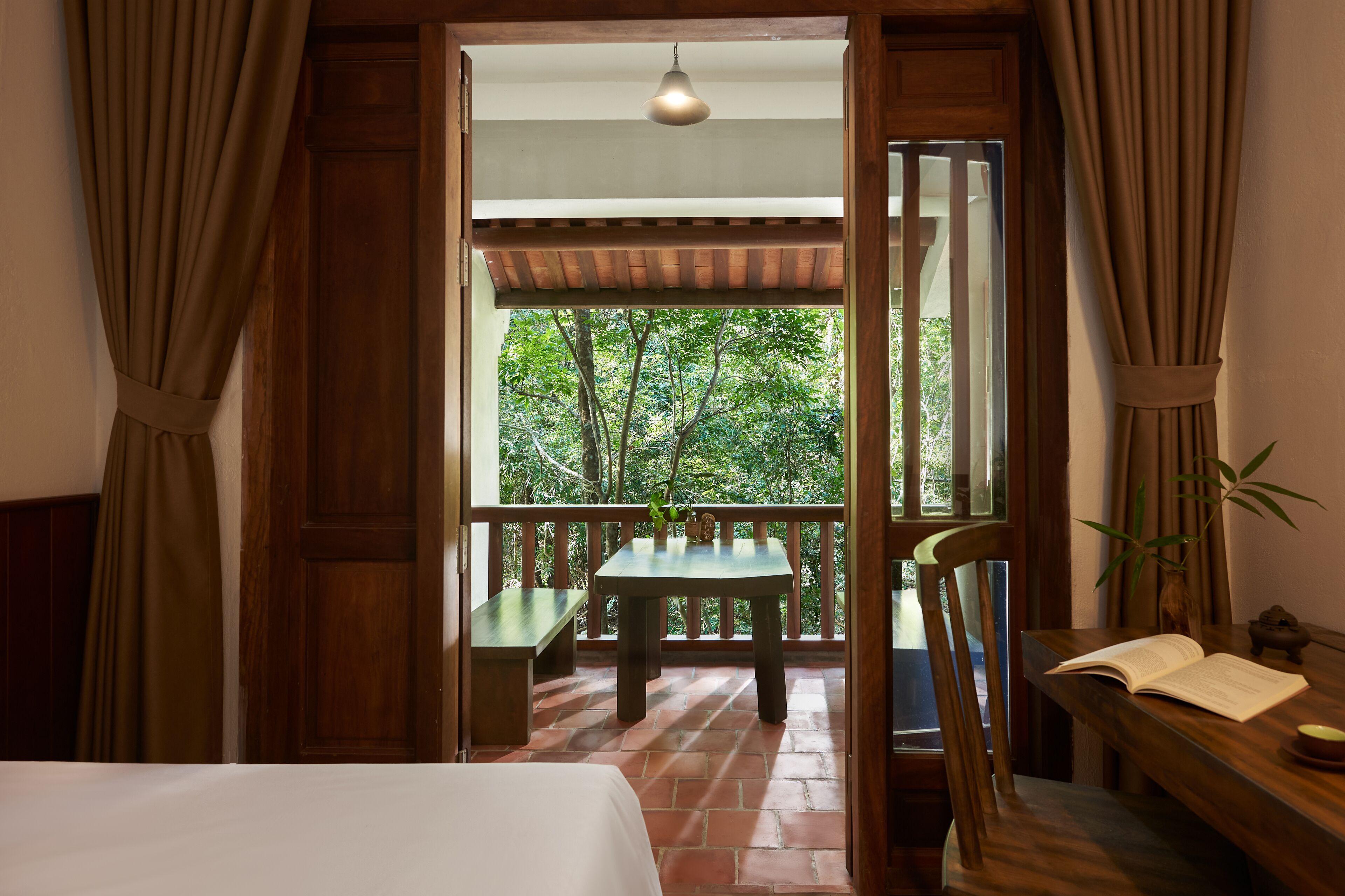 A room with a bed, a wooden desk with an open book, a bench, and a table with a plant.