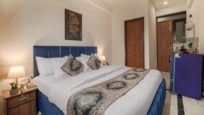 A hotel room with a bed, bedside lamps, a wooden nightstand, a blue headboard, a decorative wall hanging, a wooden door, and a purple refrigerator. - Aerostays Studio Apartments (Dera Bassi)