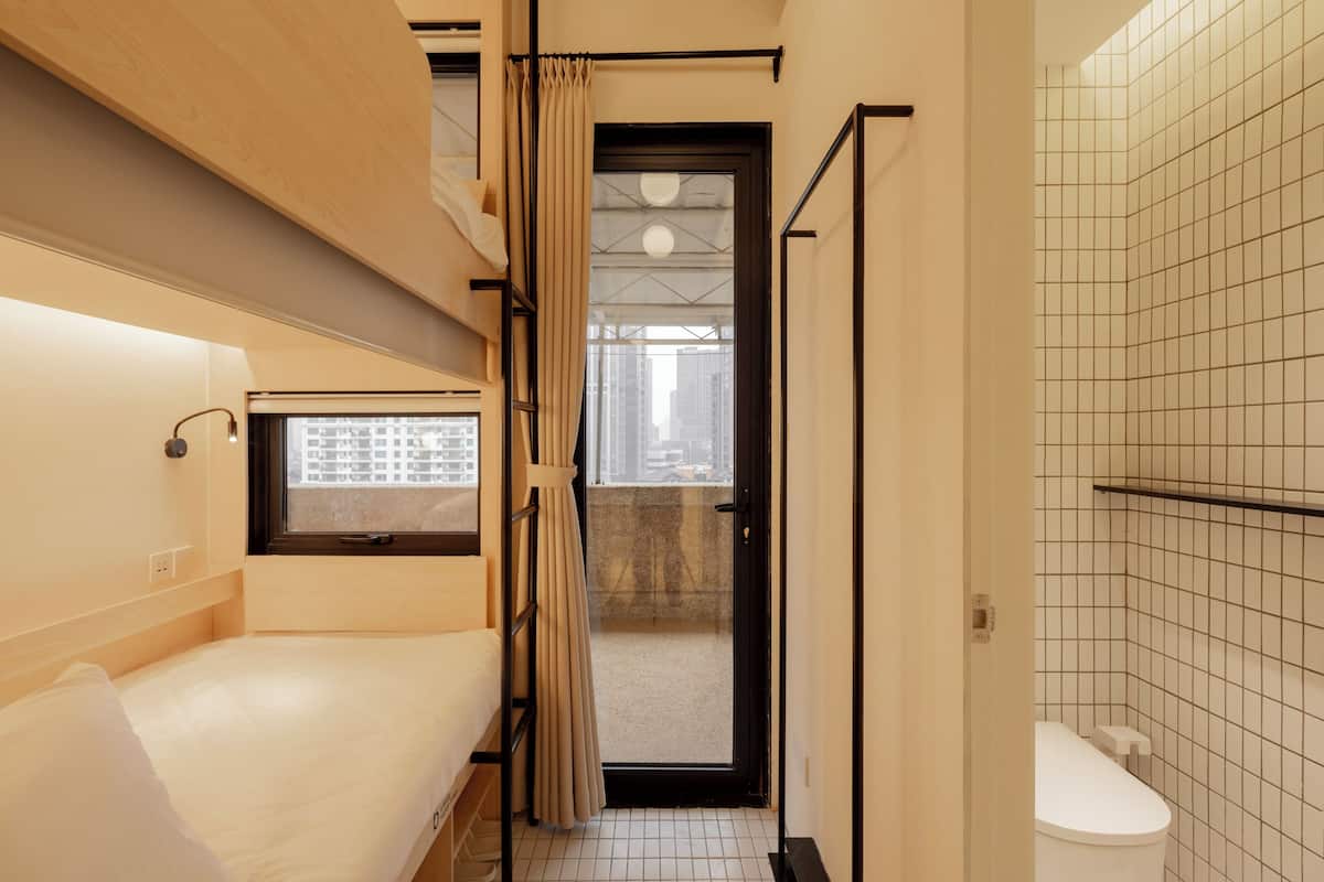 Ein kompaktes Hotelzimmer mit Etagenbett, kleinem Fenster, Dusche und Blick auf die Stadt durch die Tür.