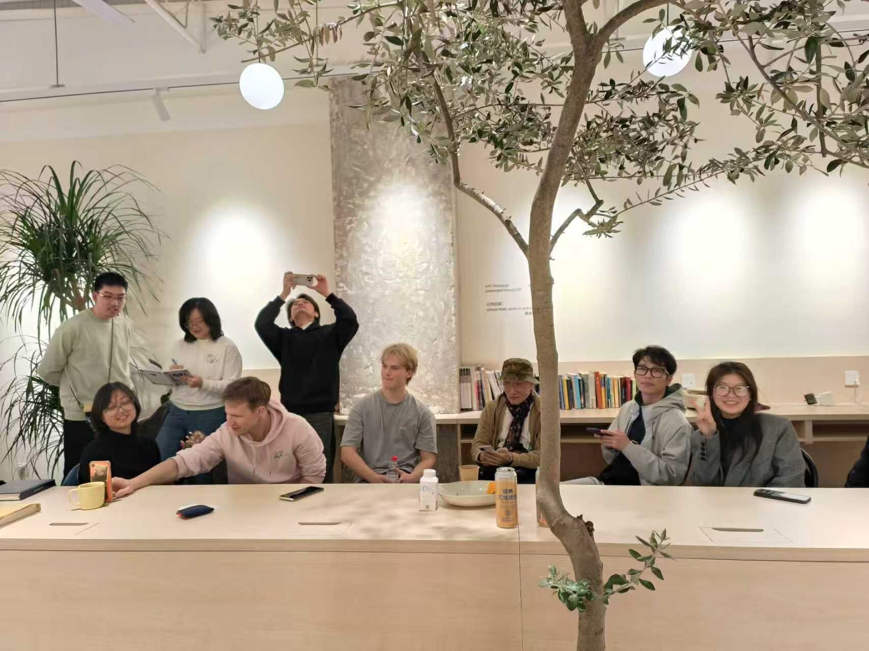A group of people in a modern indoor setting with a central table, surrounded by bookshelves and a potted plant.