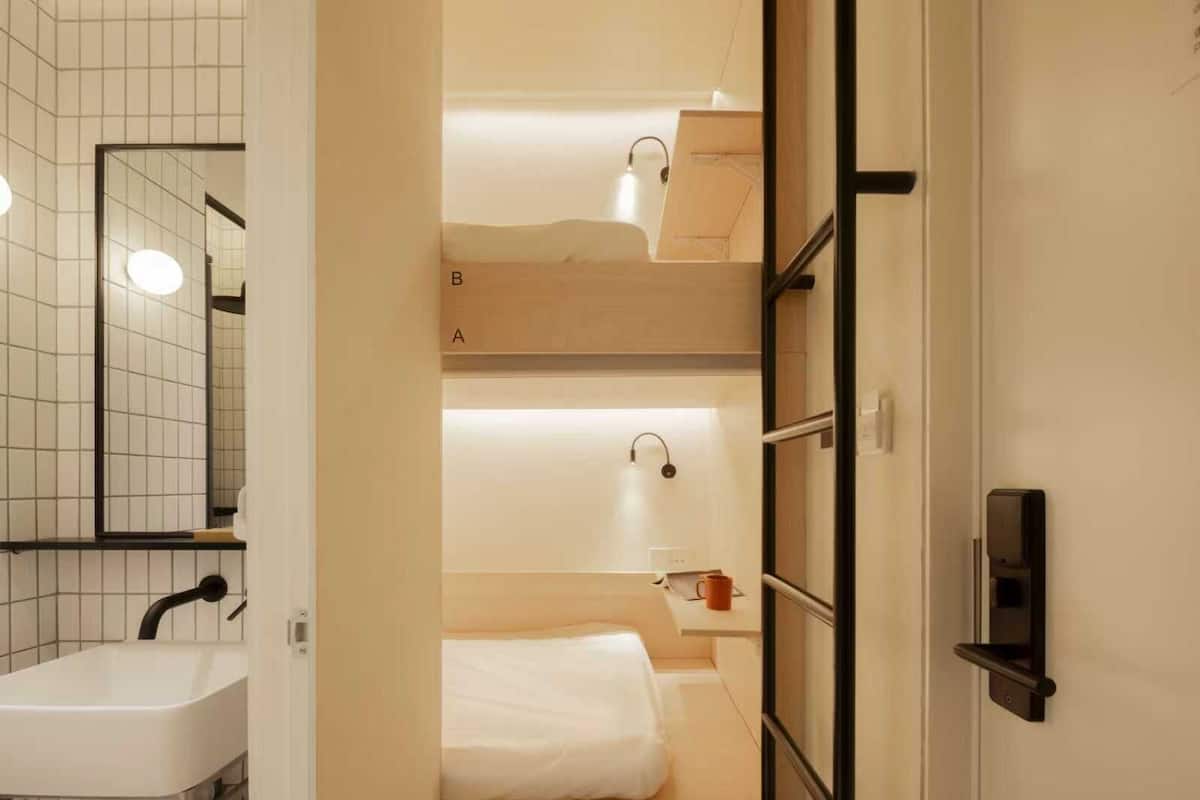 Ein kompaktes Hotelzimmer mit Bad, das ein Waschbecken, einen Spiegel und einen Handtuchhalter umfasst. Ein kleiner Tisch mit einer Tasse und einem Handtuch steht bereit. Das Zimmer verfügt über Stockbetten, die mit A und B gekennzeichnet sind.