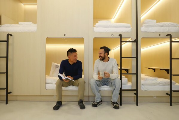 A hostel room with bunk beds labeled B, C, and D, and a sitting area with two people.