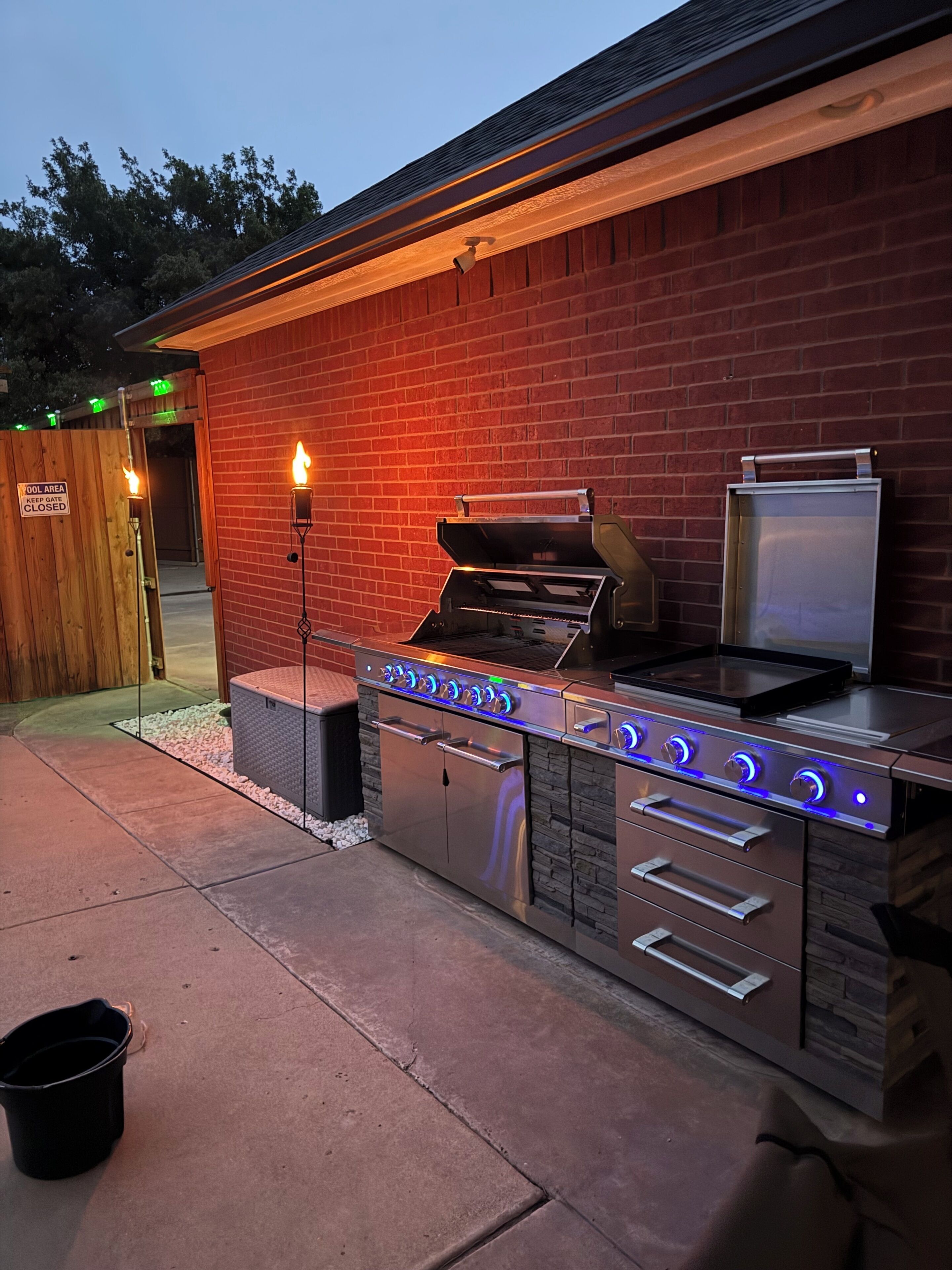 An outdoor kitchen with a grill, oven, and built-in refrigerator, set against a brick wall.