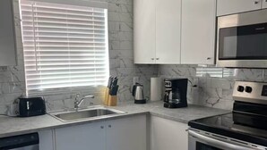 A modern kitchen with white cabinets, a stainless steel dishwasher, a built-in oven, and a window with blinds. - Beautiful Townhome Near Sanibel (Fort Myers)