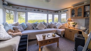 Ein gemütliches Zimmer mit einer Couch, einem Holztisch, einem Schreibtisch mit Lampe und Blick durch große Fenster.