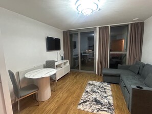Ein modernes Wohnzimmer mit einem runden Tisch, zwei Stühlen, einem grauen Sofa, einem an der Wand befestigten Fernseher und einer Glastür zum Balkon.