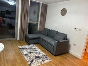 Ein modernes Wohnzimmer mit einem grauen Ecksofa, einem gemusterten Teppich, einer Wanduhr und einer Schiebeglastür, die auf einen Balkon führt.