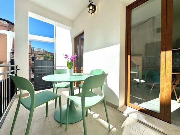 En balkon med et rundt bord, fire stole, en potteplante og udsigt til en bygning i baggrunden.