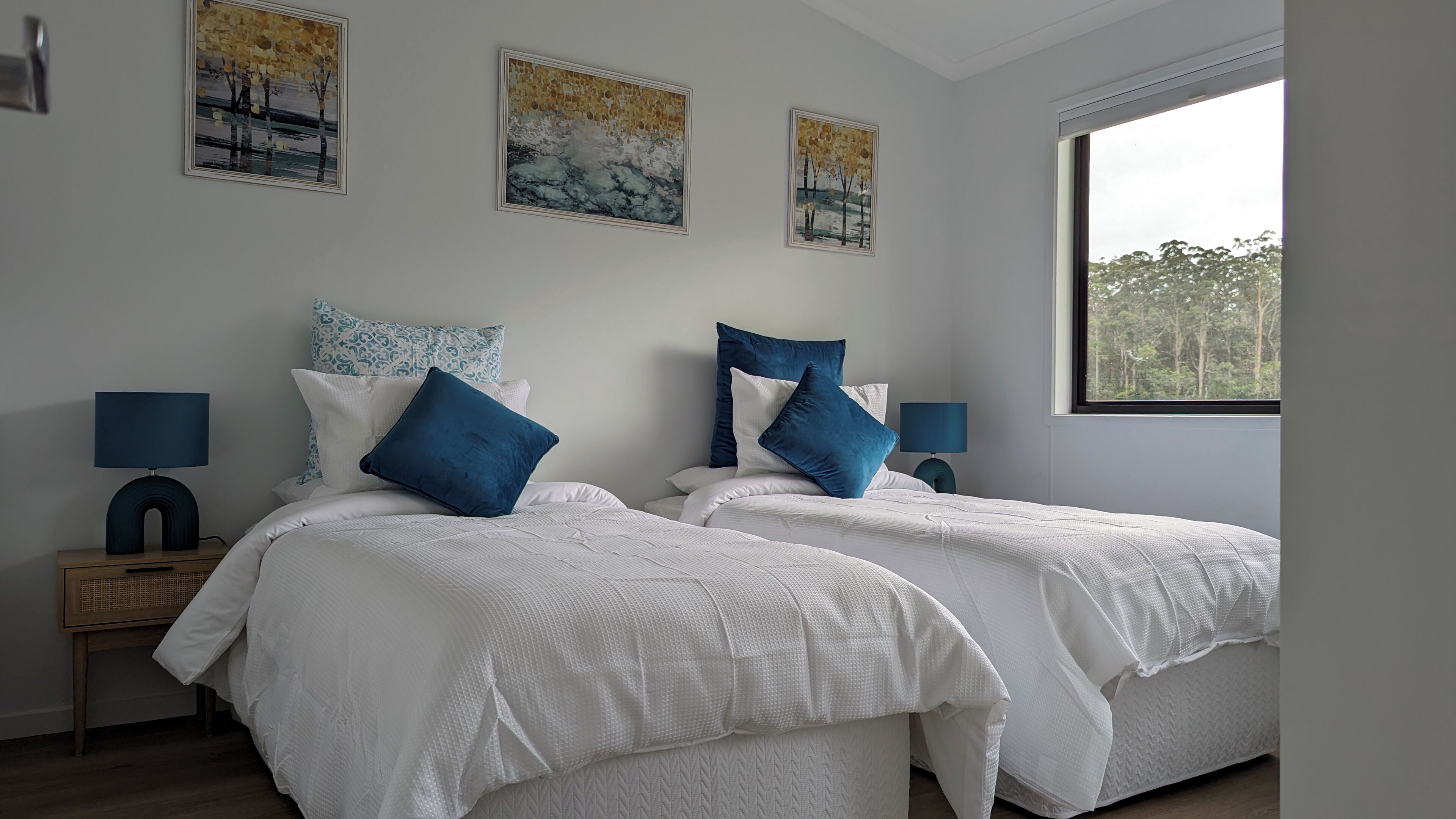 A bedroom with two beds, white bedding, blue pillows, and a nightstand with a lamp.