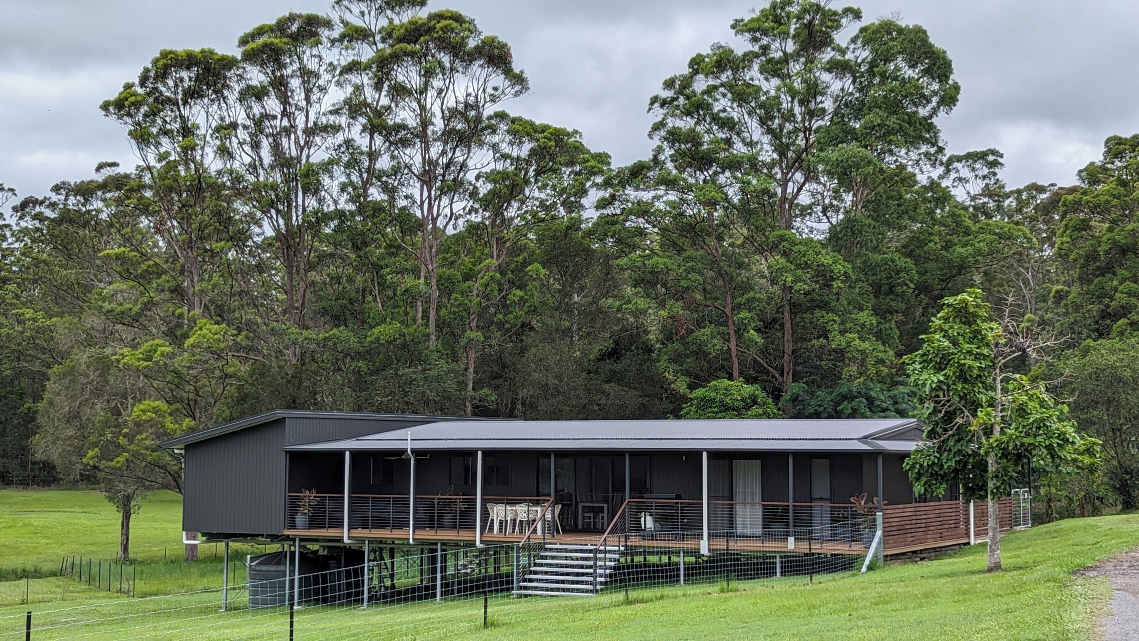 A modern house with a large deck, surrounded by a fenced yard and dense forest.