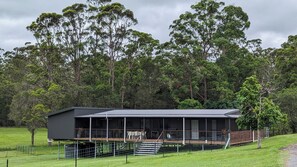 A modern house with a large deck, surrounded by a fenced yard and dense forest. - Luxury Home in Countryside Setting close to Beaches, Bushland, Restaurants/Shops (North Arm)