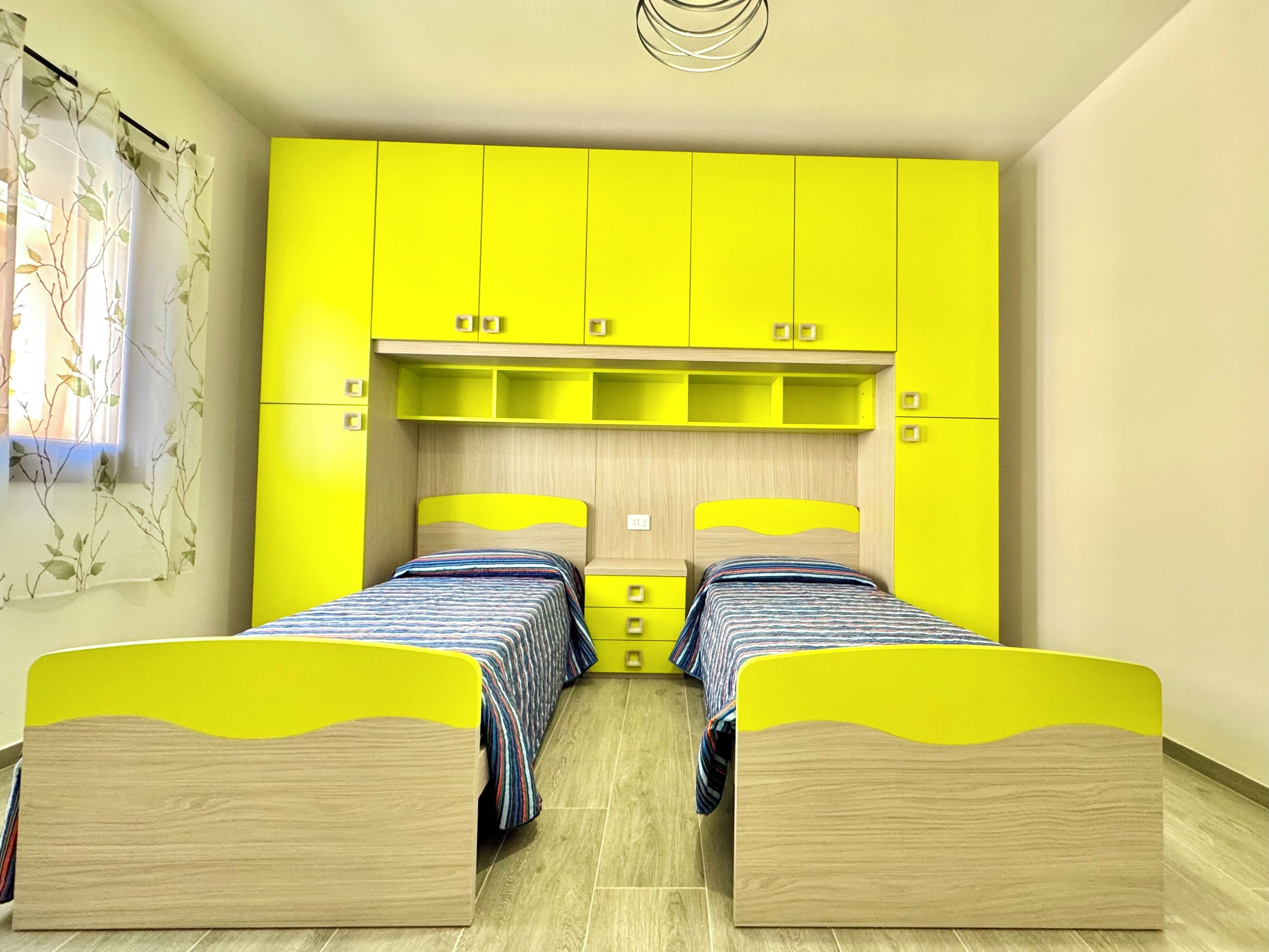 Una habitación con dos camas, armarios amarillo brillante y una ventana con una planta.
