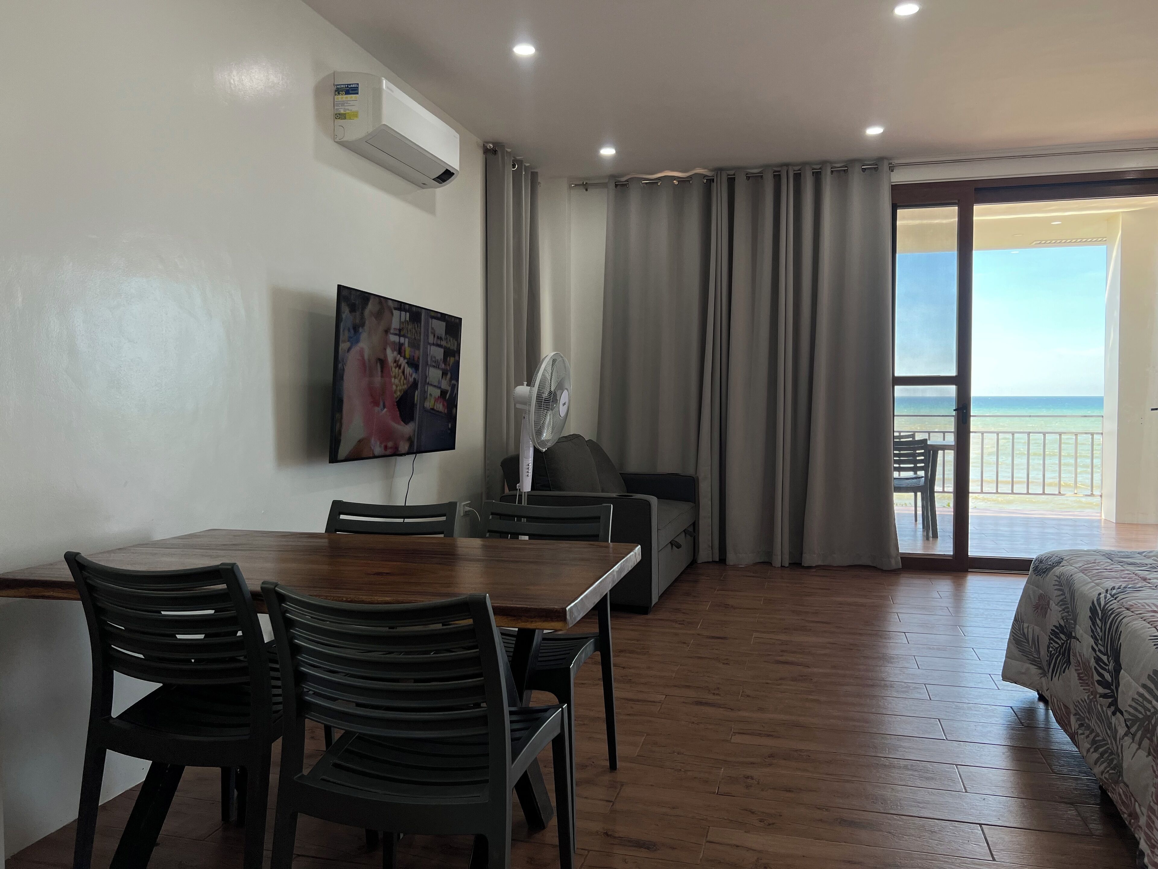 Habitación de hotel con piso de madera, una mesa de comedor con sillas, un sofá, un televisor montado en la pared y un balcón con vista al mar.