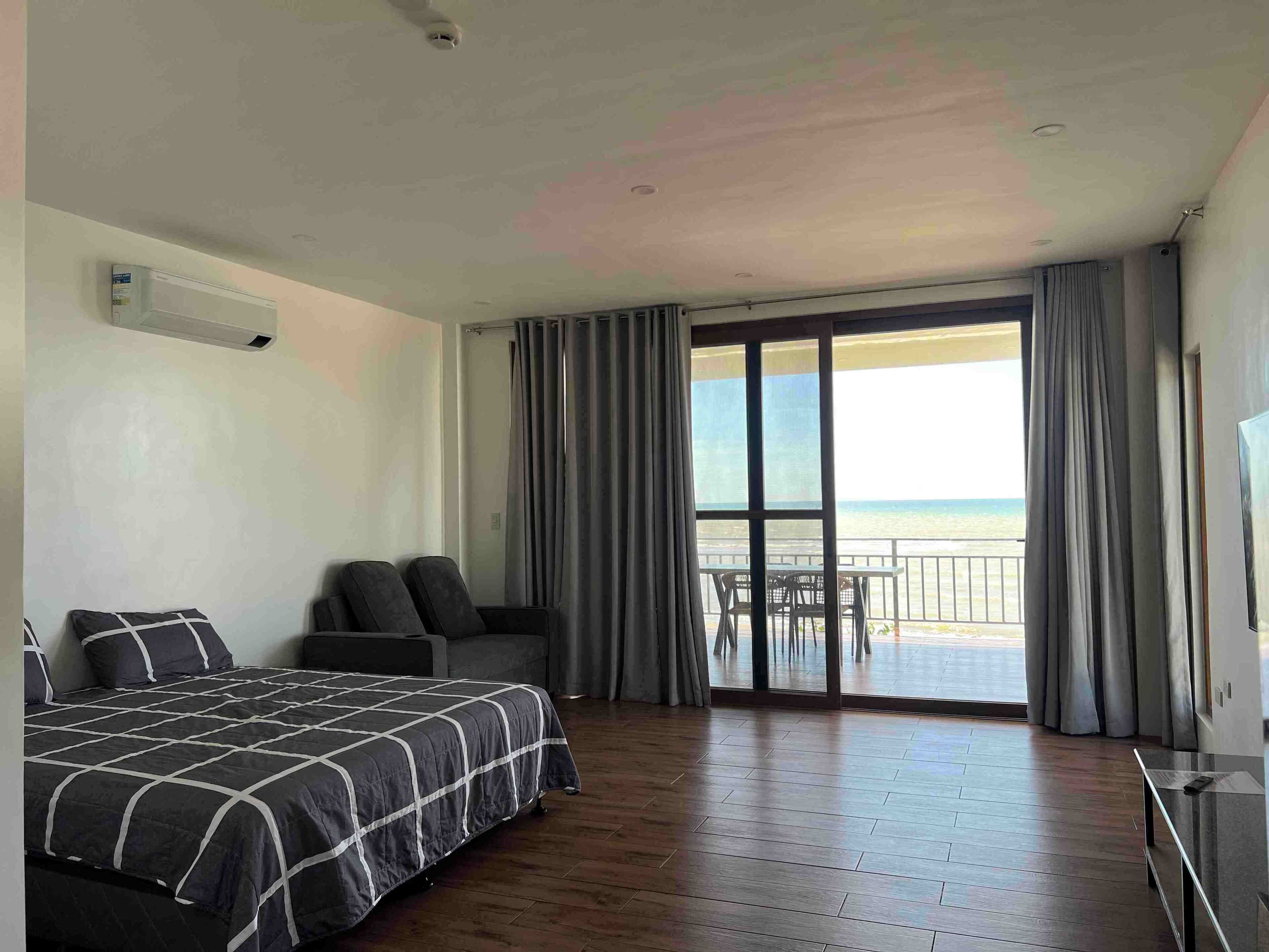 A hotel room with a bed, a seating area with chairs, a large window with a view of the sea, and a balcony with a table and chairs.