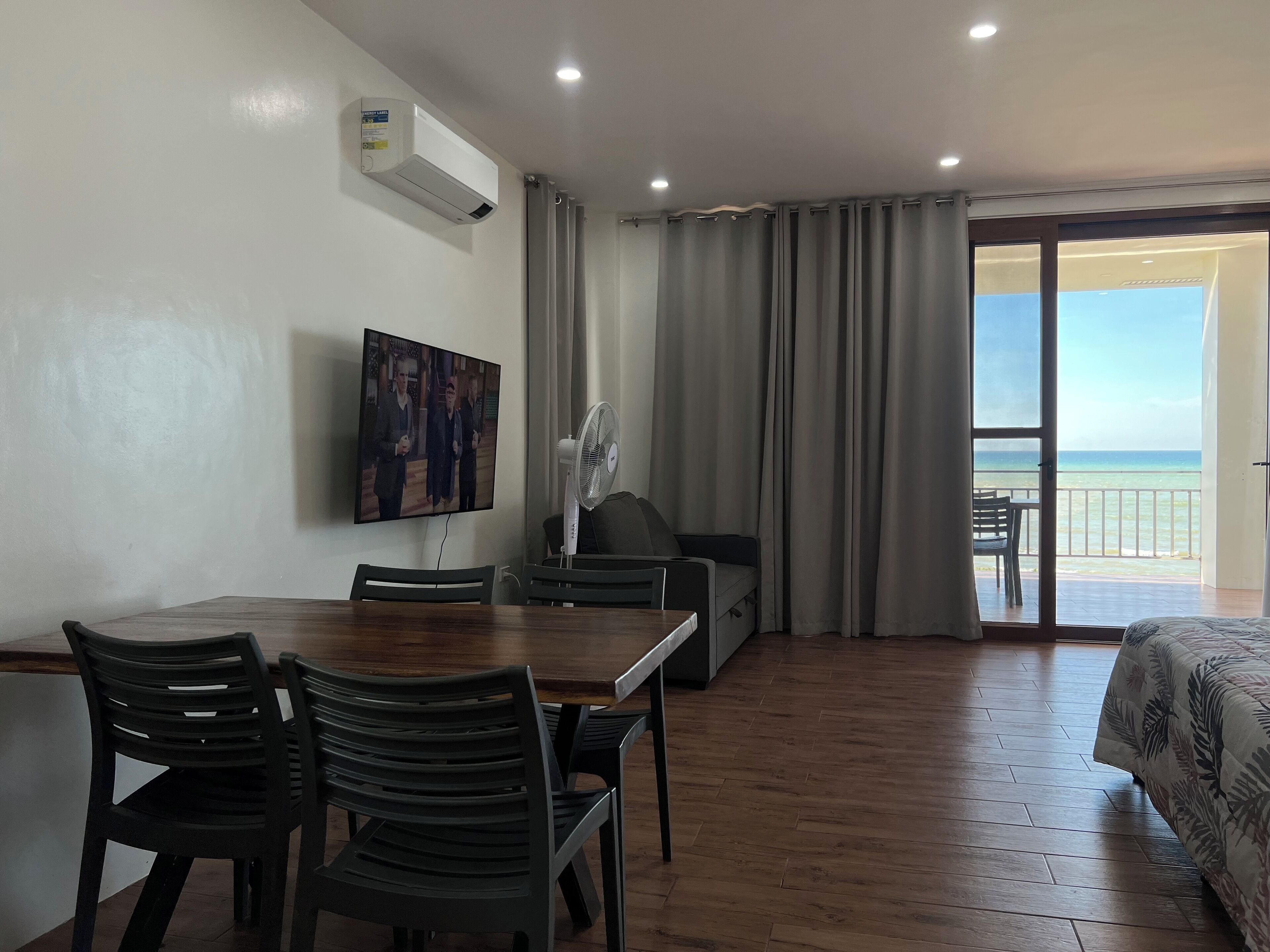 Habitación de hotel con una mesa de comedor de madera, sillas, televisor, ventilador, sofá y balcón con vista al mar.