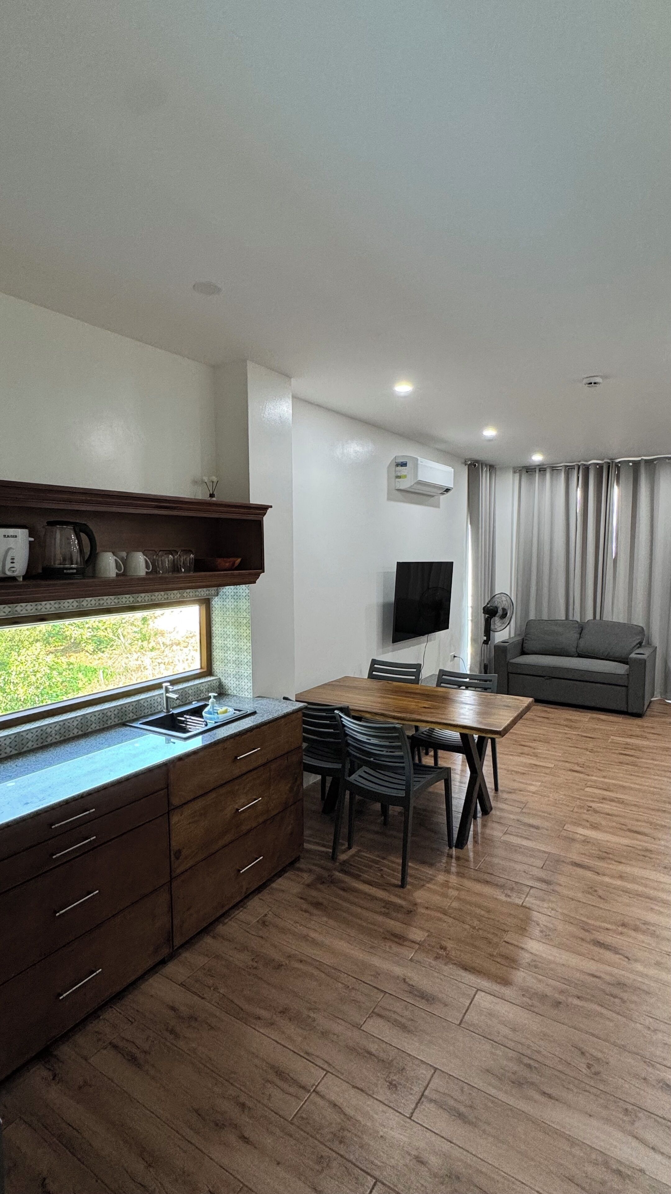 A modern kitchen with wooden cabinets, a dining area with a table and chairs, a grey sofa, and a TV mounted on the wall.