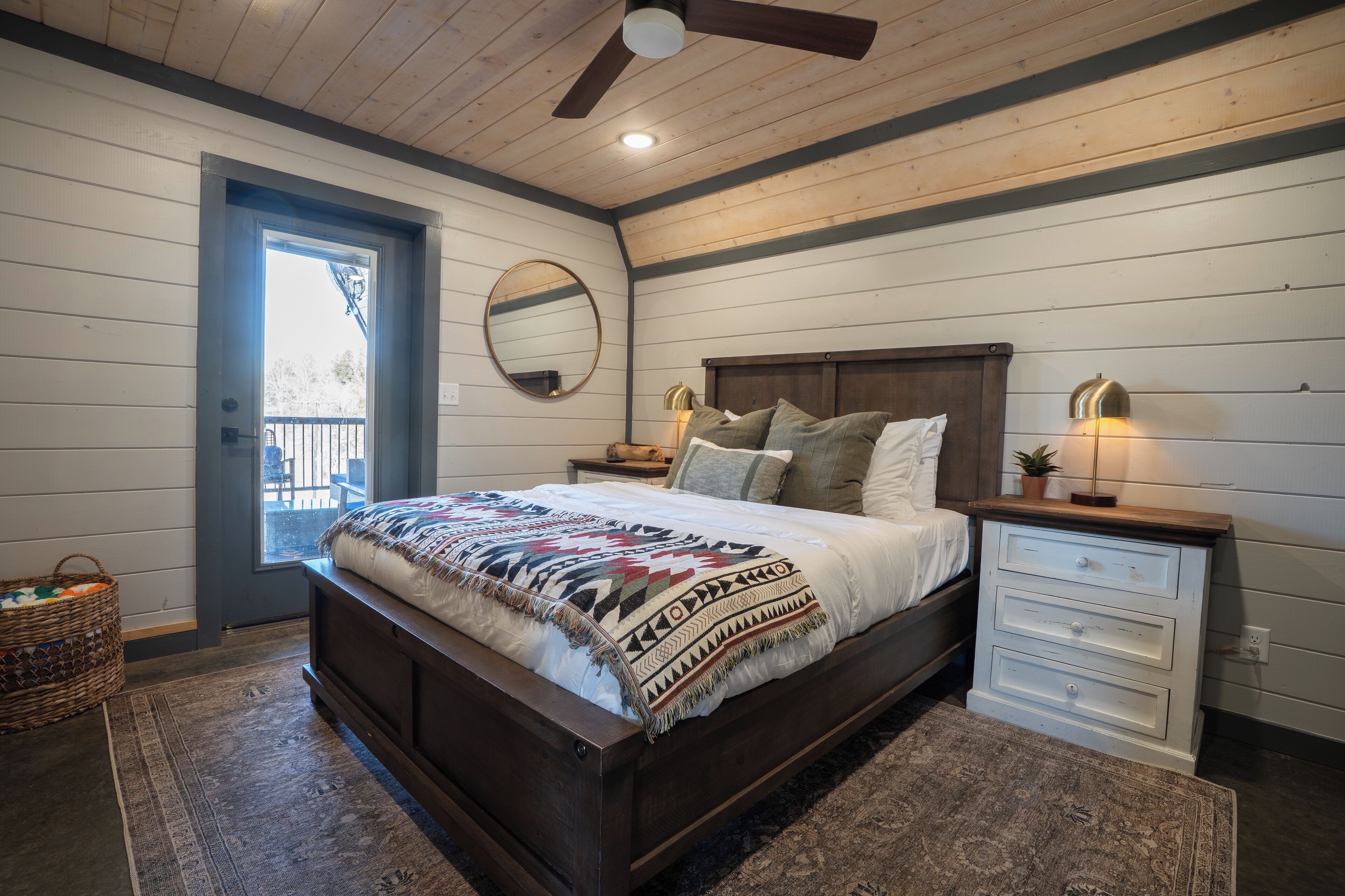 A bedroom with a wooden bed, a nightstand with drawers, a mirror, and a window with a view of the outside.