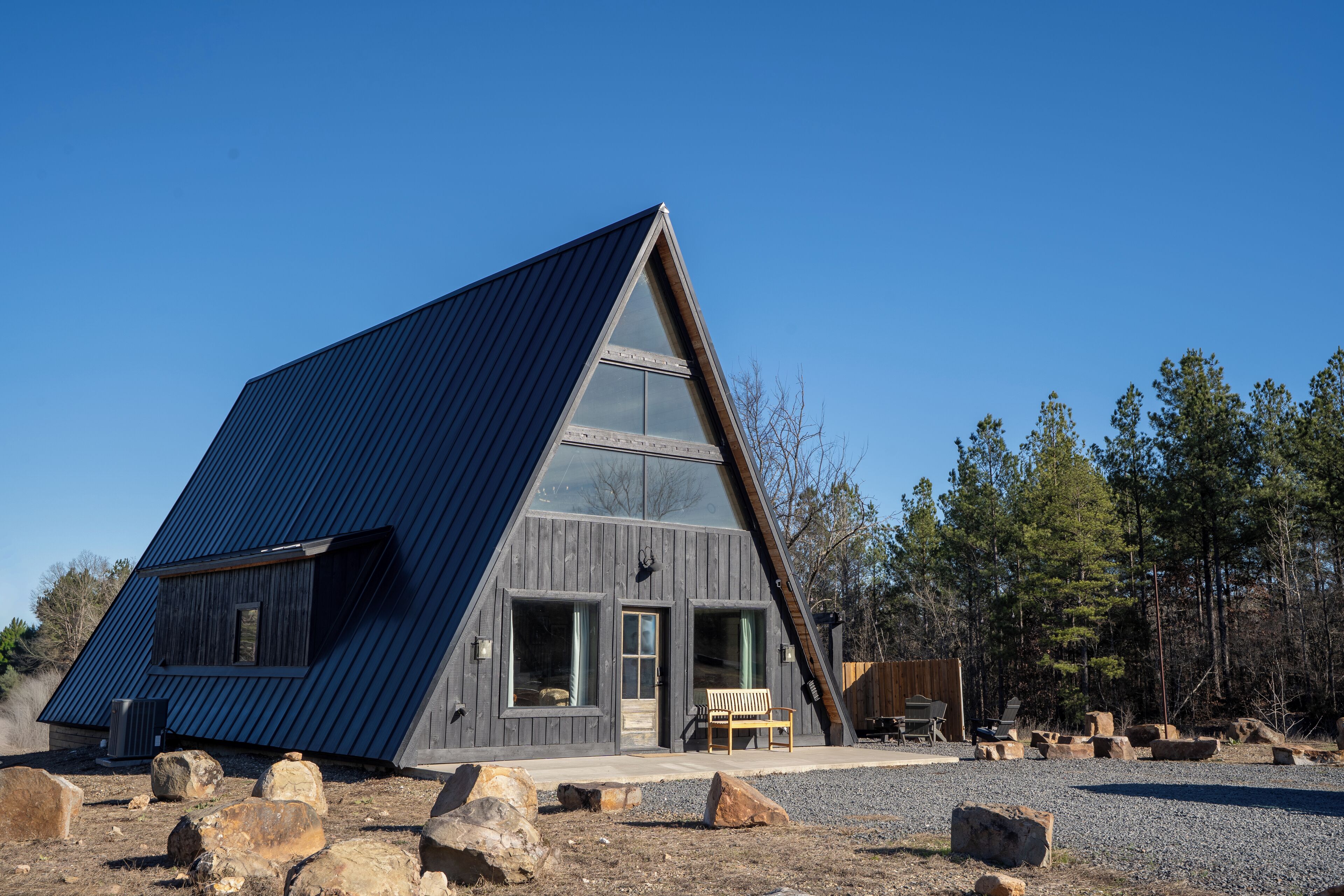 A modern A-frame cabin with large windows, a wooden deck, and a gravel ground.