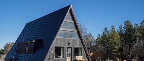 A modern A-frame cabin with large windows, a wooden deck, and a gravel ground.