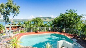 A pool with a curved edge, surrounded by a white fence, with a view of a lake and hills in the background. - Heliconia 4 & Free Buggy - Hamilton Island (Hamilton Island)