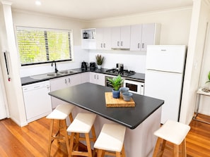 A modern kitchen with white cabinets, a black countertop, and wooden stools. - Panorama 5 - Hamilton Island & Buggy & Transfers (Hamilton Island)