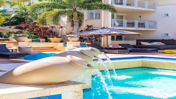 En swimmingpool med et fareskilt, liggestole og en bygning med altaner i baggrunden.