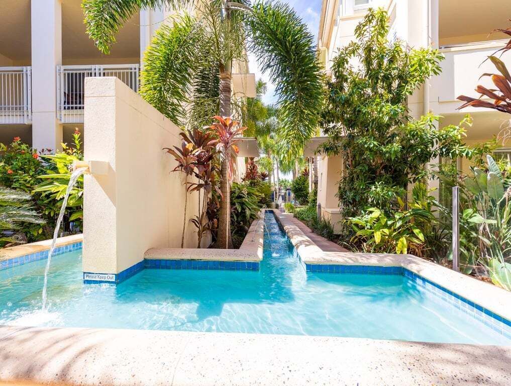 Una piscina con cascada, rodeada de exuberante follaje y un edificio de varios pisos.