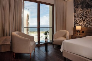 A hotel room with a large window, two armchairs, a small table with a plant, a bed, and a decorative wall piece.