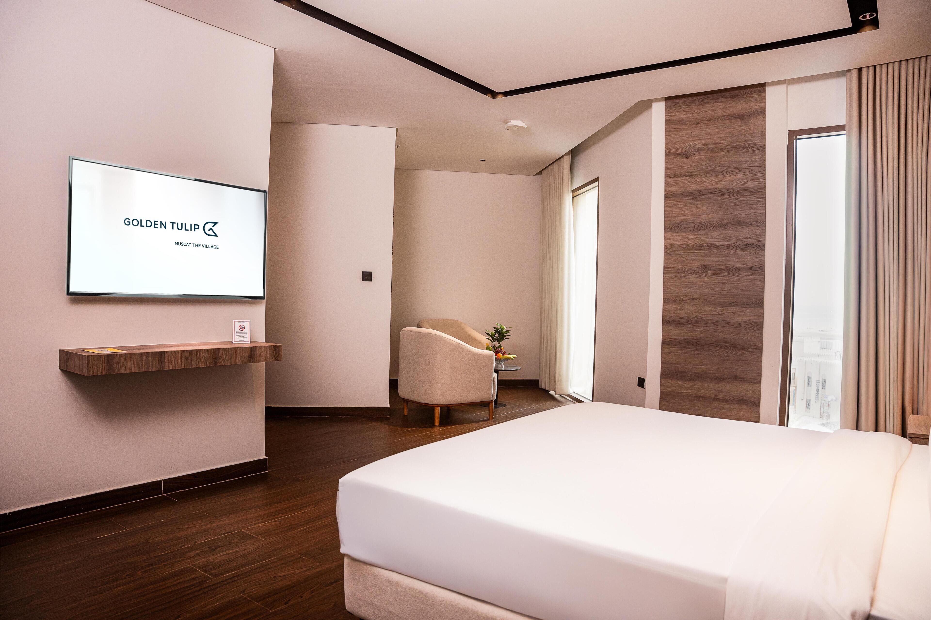 A hotel room with a bed, a TV mounted on the wall, a wooden shelf, a chair, and a window with curtains.