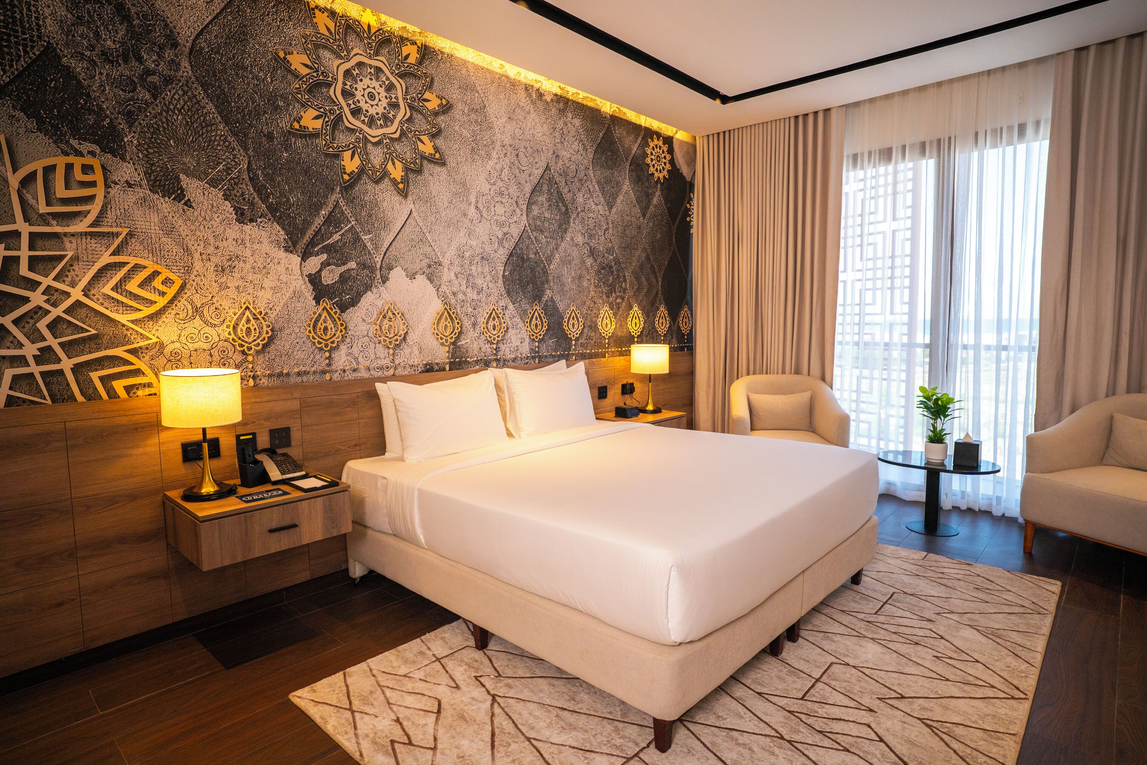 Une chambre d’hôtel avec un grand lit, deux tables de chevet, une chaise, une petite table avec une plante et un mur décoratif.