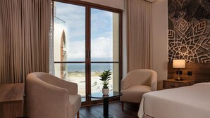 A hotel room with a large sliding glass door, two armchairs, a small round table with a plant, a bed, and a nightstand with a lamp. - Golden Tulip Muscat The Village (Seeb)