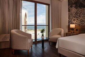Una habitación de hotel con un gran ventanal corredero, dos sillones, una mesita redonda con una planta, una cama y una mesita de noche con una lámpara.