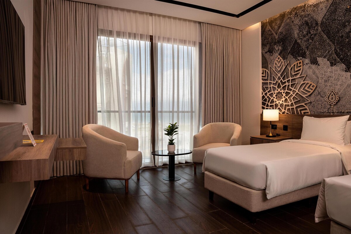 Ein Hotelzimmer mit einem Bett, zwei Sesseln, einem kleinen Tisch mit einer Pflanze, einem Nachttisch mit einer Lampe und einem großen Fenster mit transparenten Vorhängen.