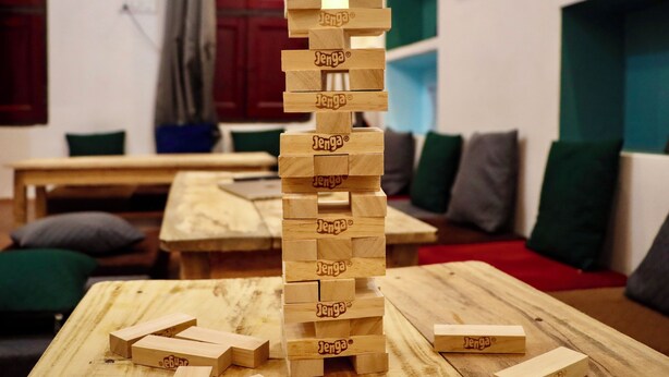 Une tour de Jenga est posée sur une table en bois dans une pièce où se trouvent un canapé avec des coussins verts et gris, ainsi qu’une couverture rouge sur le sol.