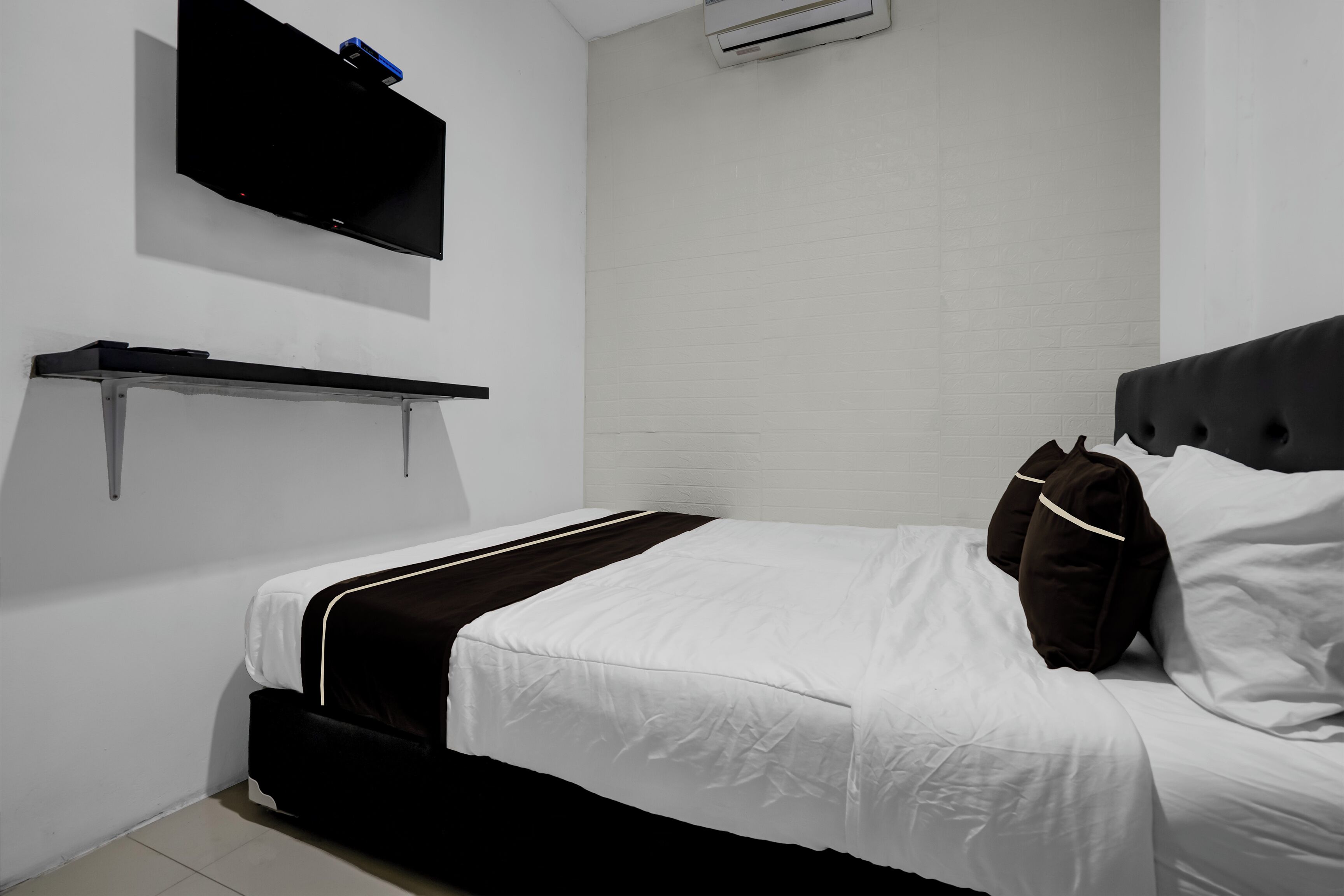 A neatly made bed with white and black bedding, a mounted flat-screen TV, and a wall-mounted shelf.
