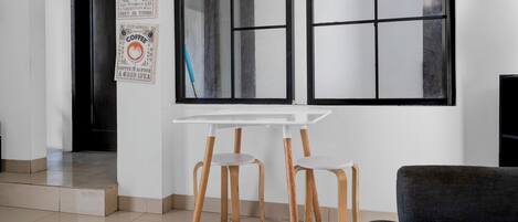 Ein modernes Zimmer mit einem weißen Tisch, zwei hölzernen Stühlen, einem dunklen Sofa und einem großen Fenster mit Ausblick.