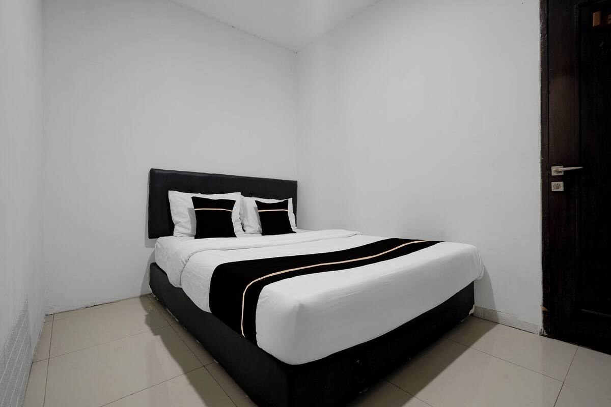 Un lit bien fait, avec une tête de lit noire et du linge de lit blanc, dans une pièce minimaliste aux murs blancs et au sol carrelé.