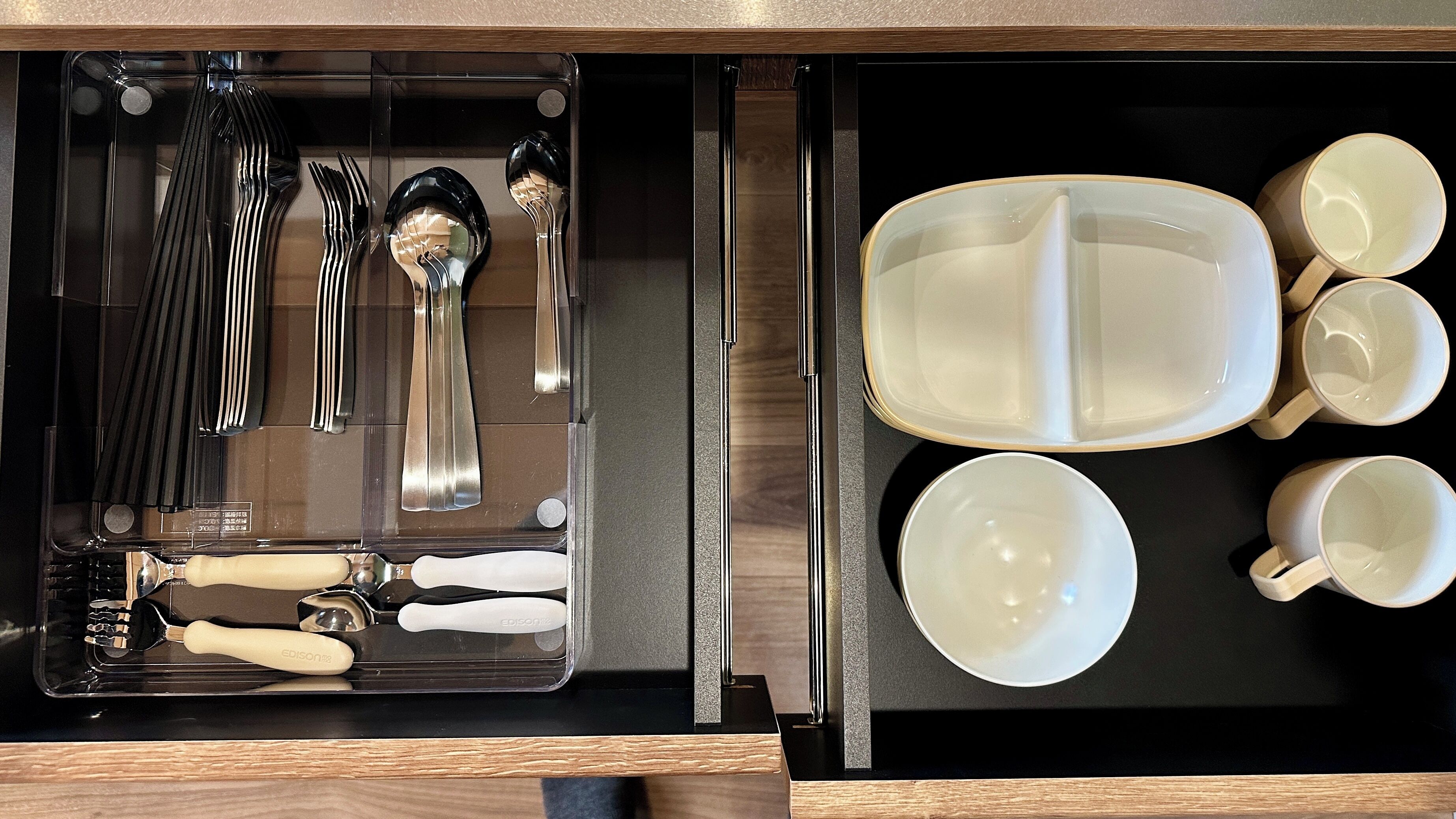 A drawer containing a set of silverware, including forks, spoons, and a knife, and a set of white ceramic cups and bowls.